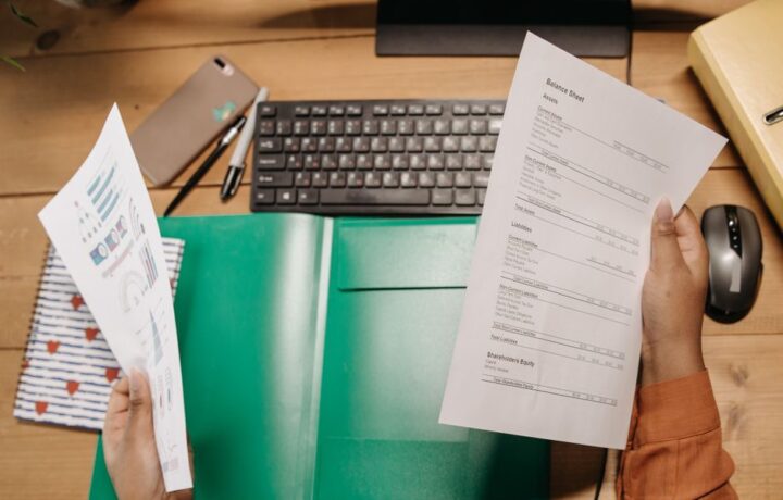 Hands holding and reviewing balance sheets over a desk with a keyboard and stationery.