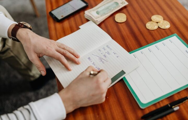 Hands writing financial calculations on notebook with money and coins on table.
