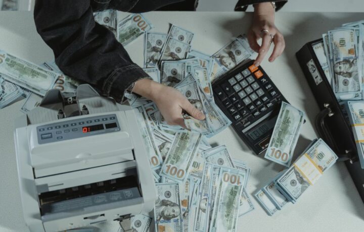 Hands counting 100 dollar bills using a calculator and money counter on a table.